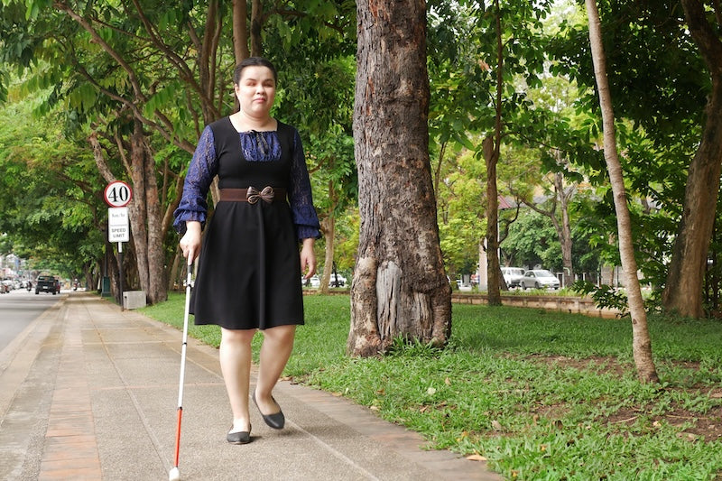 A blind woman walks confidently with a can down a wooded sidewalk.